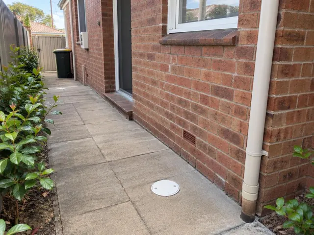 Dry paved side passage beside a brick house with a round drain cover in Arncliffe